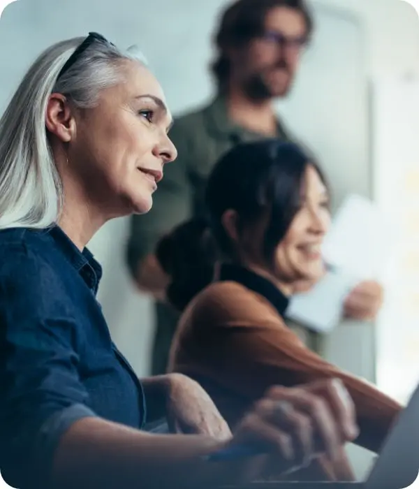 groupe de coworkers attentifs et souriants en pleine réunion ou session de travail collaboratif, avec une femme aux cheveux gris au premier plan, concentrée devant un ordinateur portable