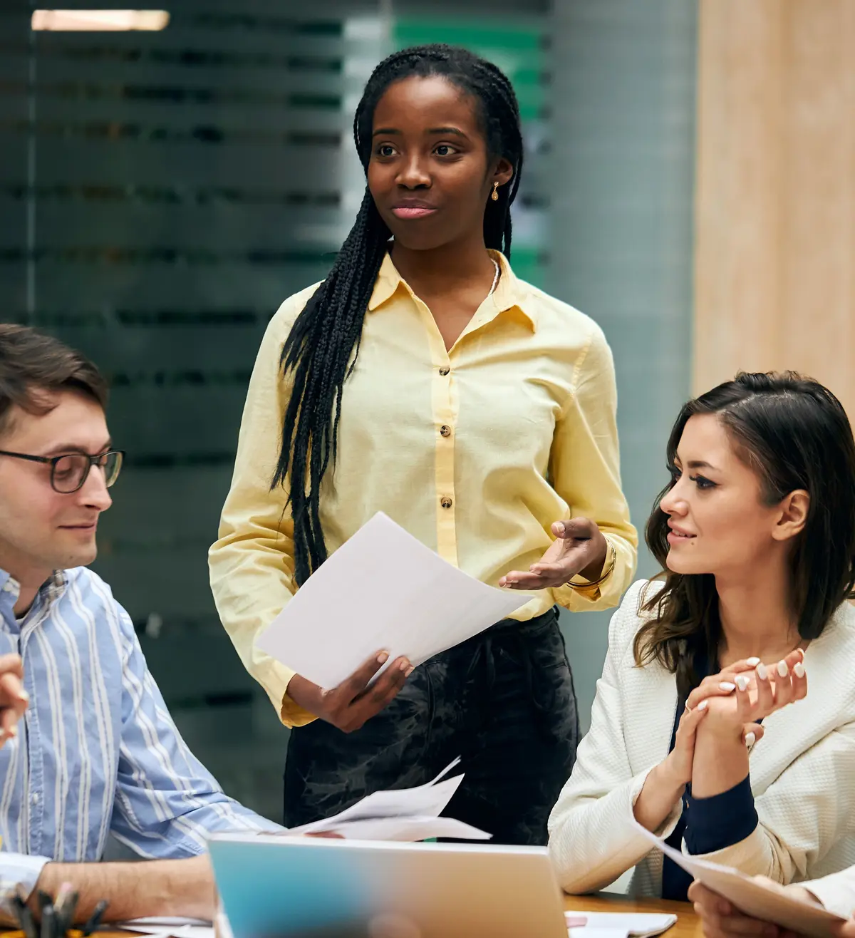 jeune femme debout tenant des feuilles, prenant la parole lors d’une réunion d’équipe en entreprise, entourée de collègues attentifs